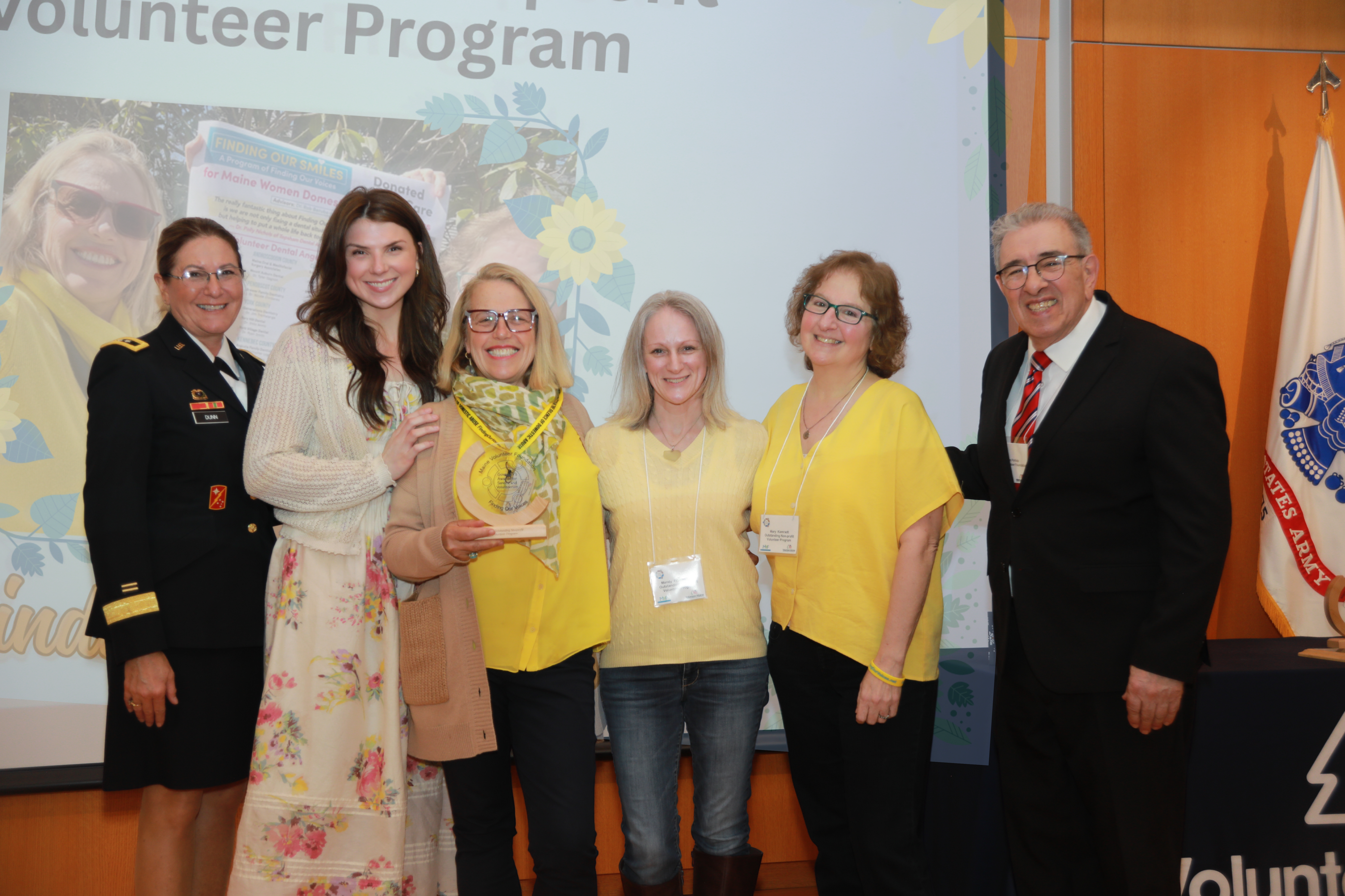 Two female representatives in yellow for Finding Our Smiles stand in between Major General Diane Dunn and Maine Volunteer Foundation President John Portela