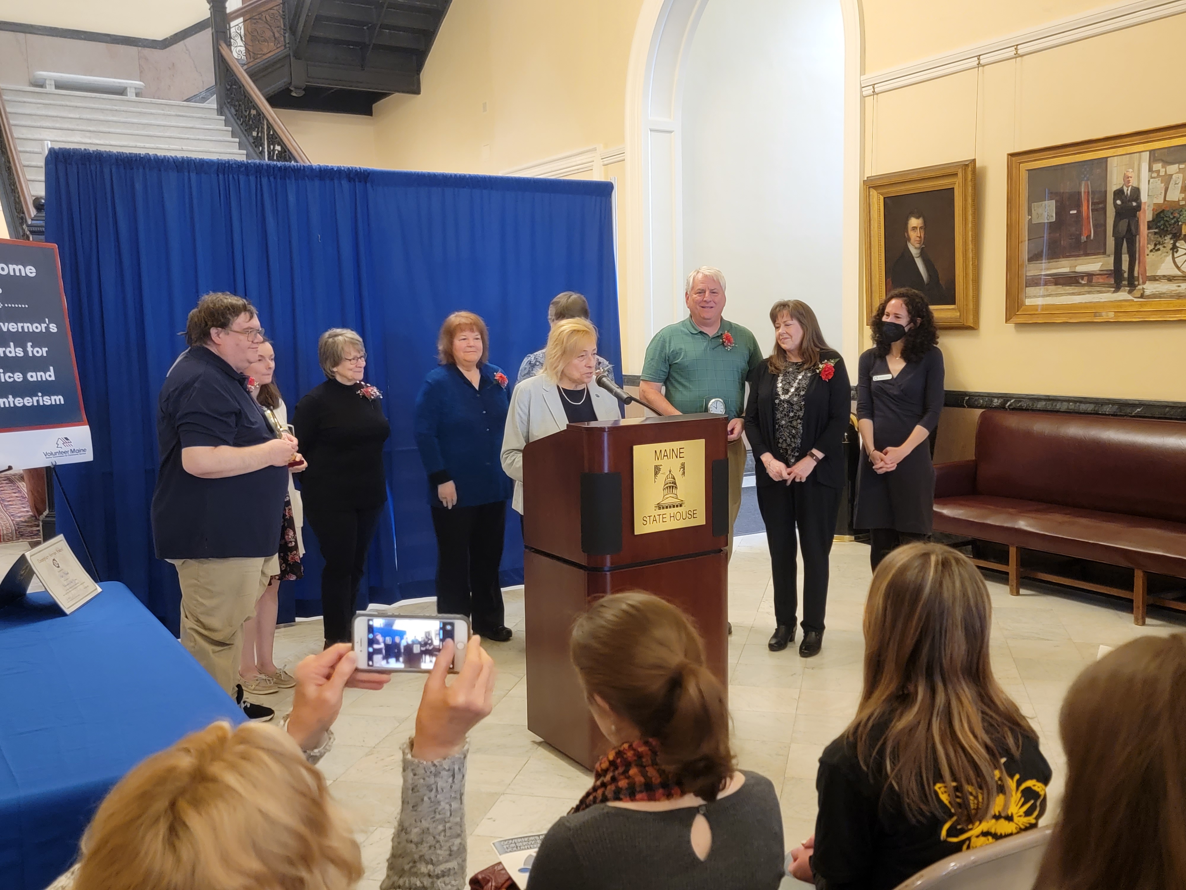 Gov. Janet Mills speaks to a crowd while at a podium with award winners behind her