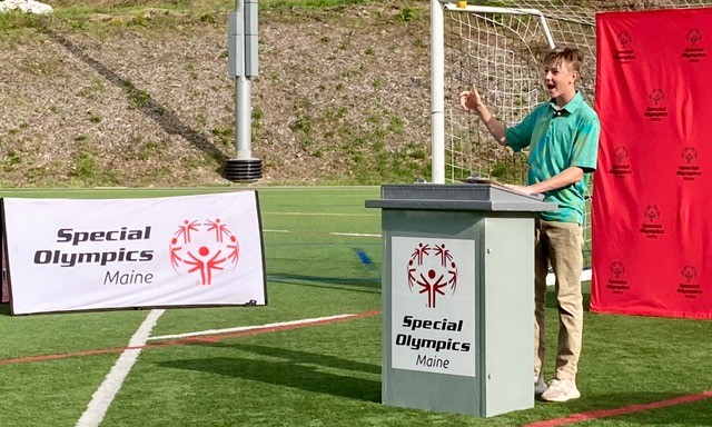 Photo of an individual speaking at an event on an athletic field on a sunny day while at a podium