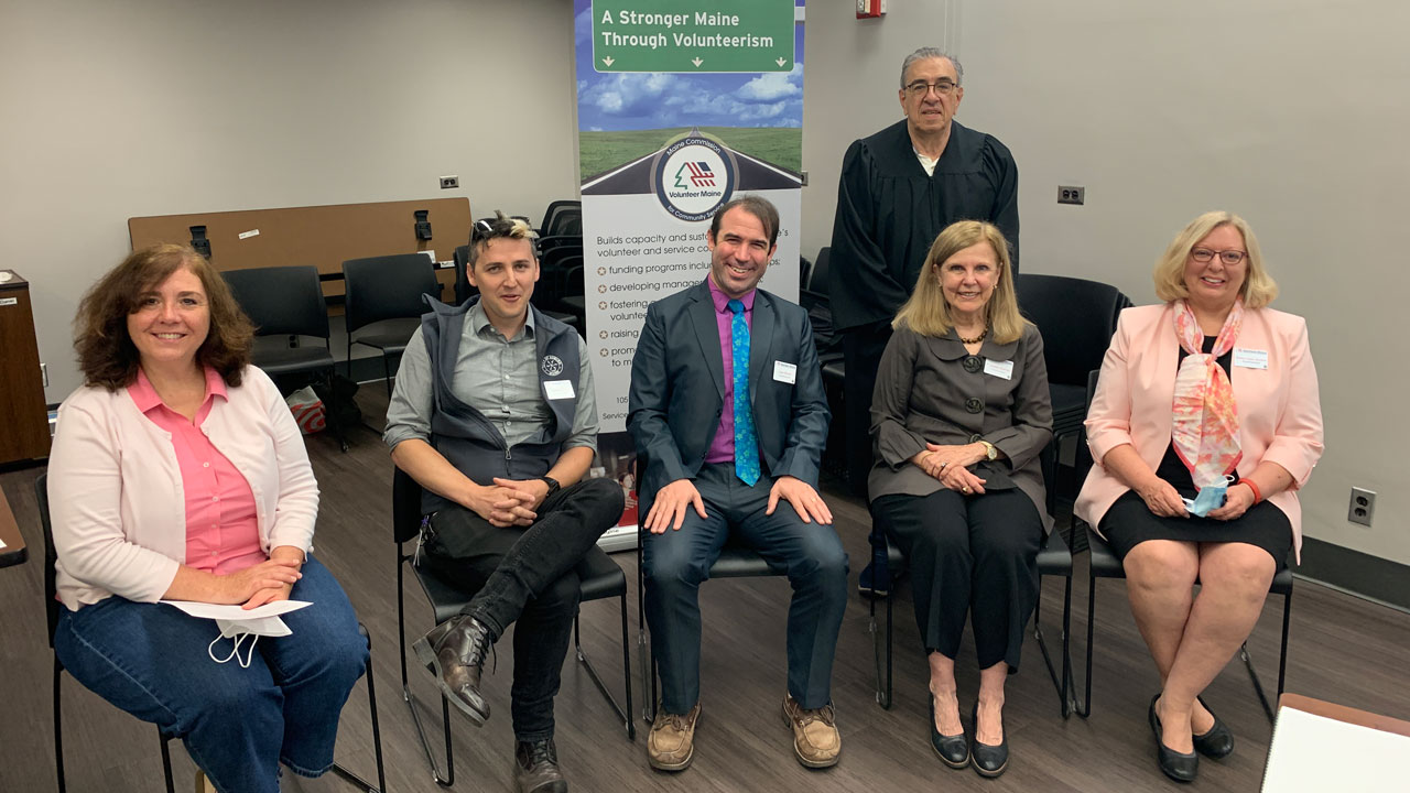 Six individuals pose for a photo in a conference room arranged five seated side by side in the front and one standing in the back