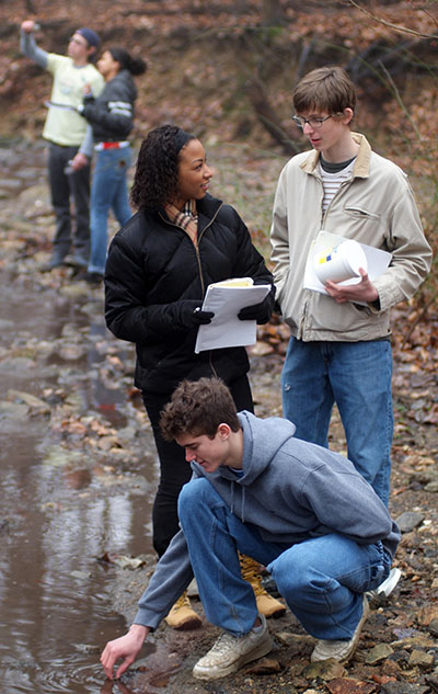 five students collect water samples by a lake and discuss findings on a cloudy day