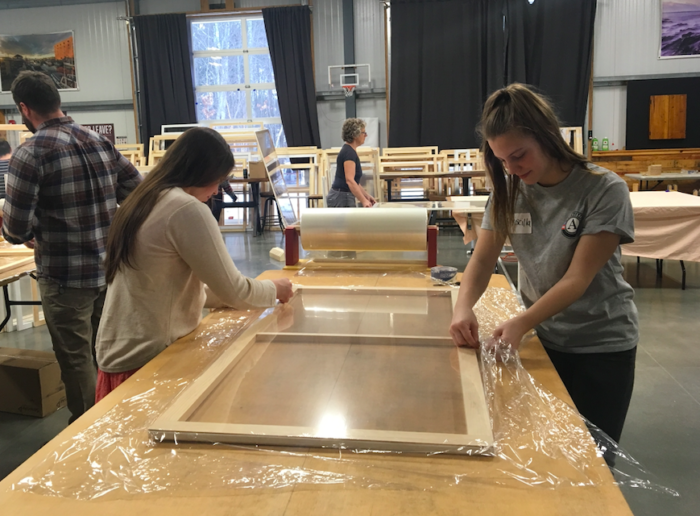Two individuals in a workshop add plastic wrap to a window insert for a weatherization project