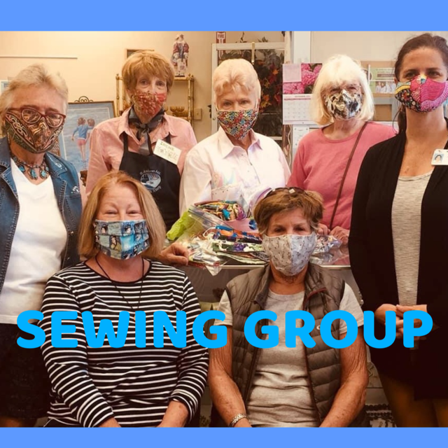 Photo of seven individuals, five standing behind two sitting, wearing face masks in a community room posing for a photo with text overlaid that reads sewing group